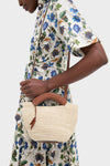 Person wearing an elegant, floral dress holds the Natural Fique Small Shopper, a classic, high-end woven straw tote with leather handles and tassel, embodying effortless style.