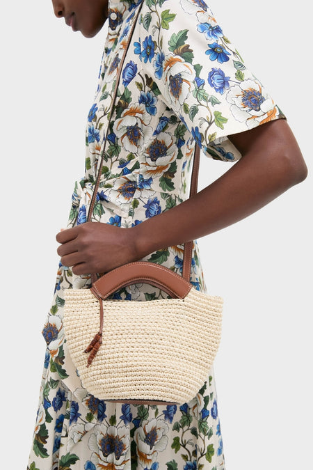 Person wearing an elegant, floral dress holds the Natural Fique Small Shopper, a classic, high-end woven straw tote with leather handles and tassel, embodying effortless style.