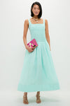 Woman in a mint green linen organza midi dress with a smocked bodice and full skirt, accessorized with gold jewelry and holding a fuchsia-pink clutch.