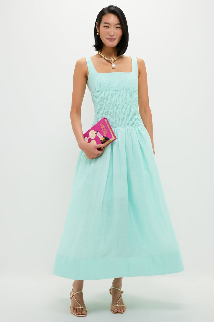 Woman in a mint green linen organza midi dress with a smocked bodice and full skirt, accessorized with gold jewelry and holding a fuchsia-pink clutch.