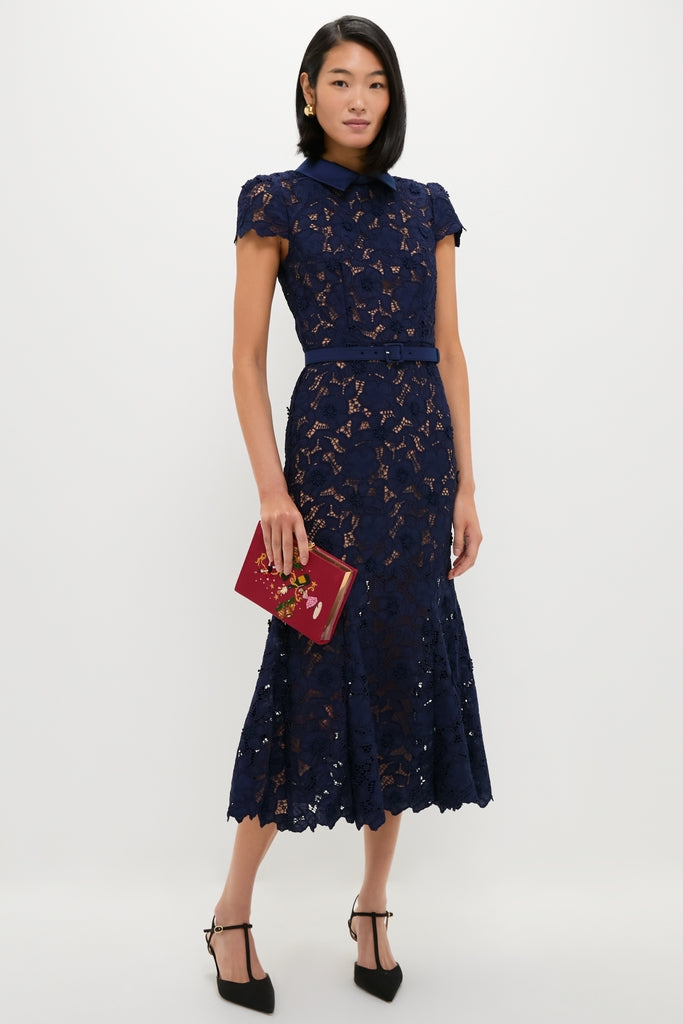 Woman in a classic navy Guipure Lace Midi Dress with cap sleeves and a pointed collar, holding a red clutch, exuding effortless elegance in high-end fashion.