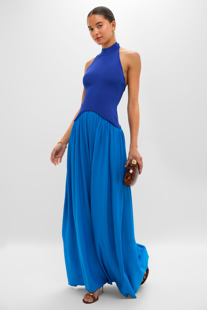 Young woman wearing the classic Cosmic Blue Soglio Halter Maxi Dress. Effortlessly styled with gold hoops, a tortoiseshell clutch, and brown strappy sandals for a high-end, fun look.