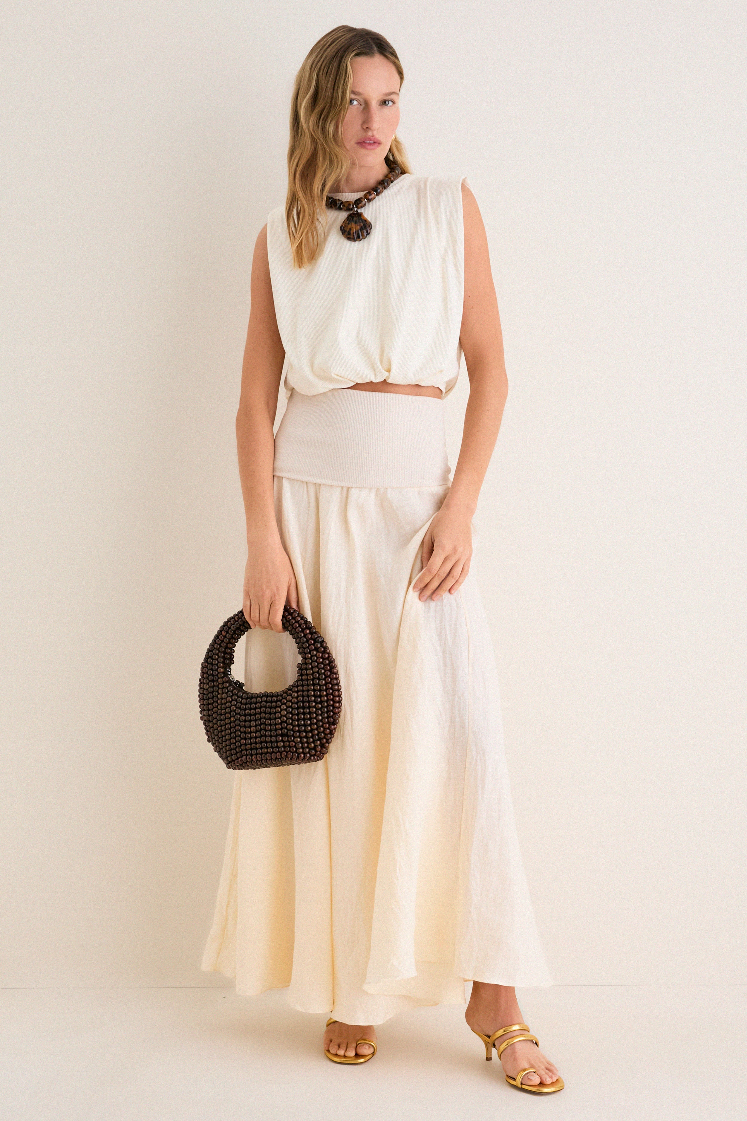 Model in ivory crop top and maxi skirt showcasing Gold Modia Sandals; classic, metallic gold with textured straps, embodying effortless high-end style, paired with a chic, beaded handbag.