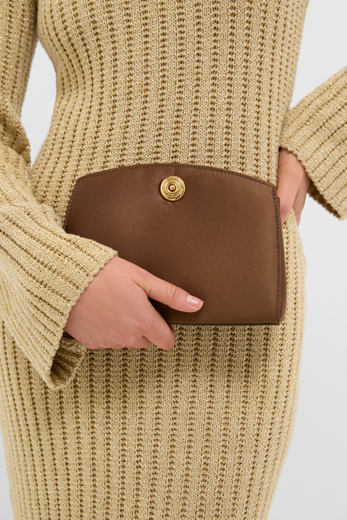Person holding a classic, high-end Brown Satin Pushlock Clutch with a curved flap and gold-toned clasp, complementing their ribbed knit attire for an effortless, fun style.