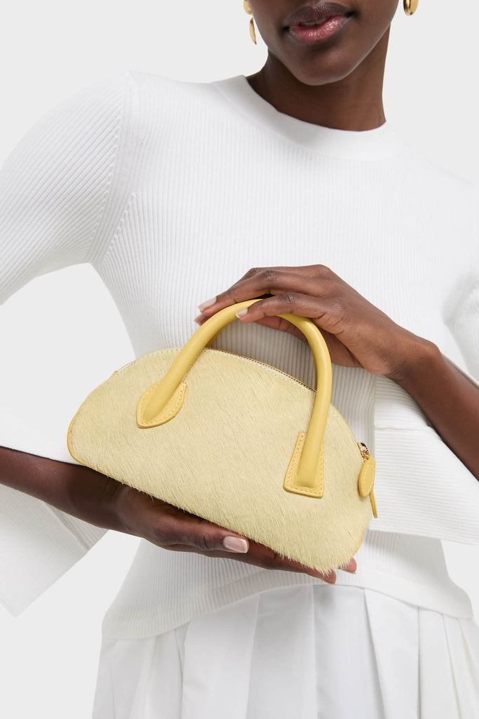 Woman holding the Butter EW Dome Clutch, a high-end, half-moon-shaped handbag in pale yellow hair-on-hide, featuring classic smooth leather handles for an effortless, fun style.