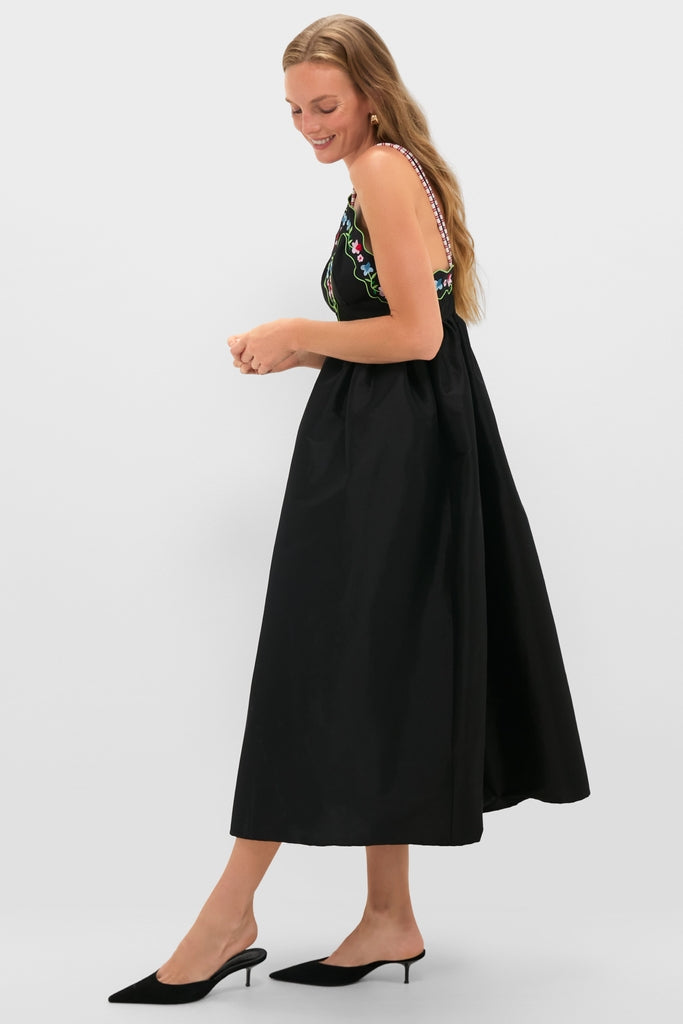 Young woman in profile wearing the Black Selene Dress, a classic, ankle-length black dress with colorful floral embroidery and contrasting straps, paired with effortless black kitten-heel mules.