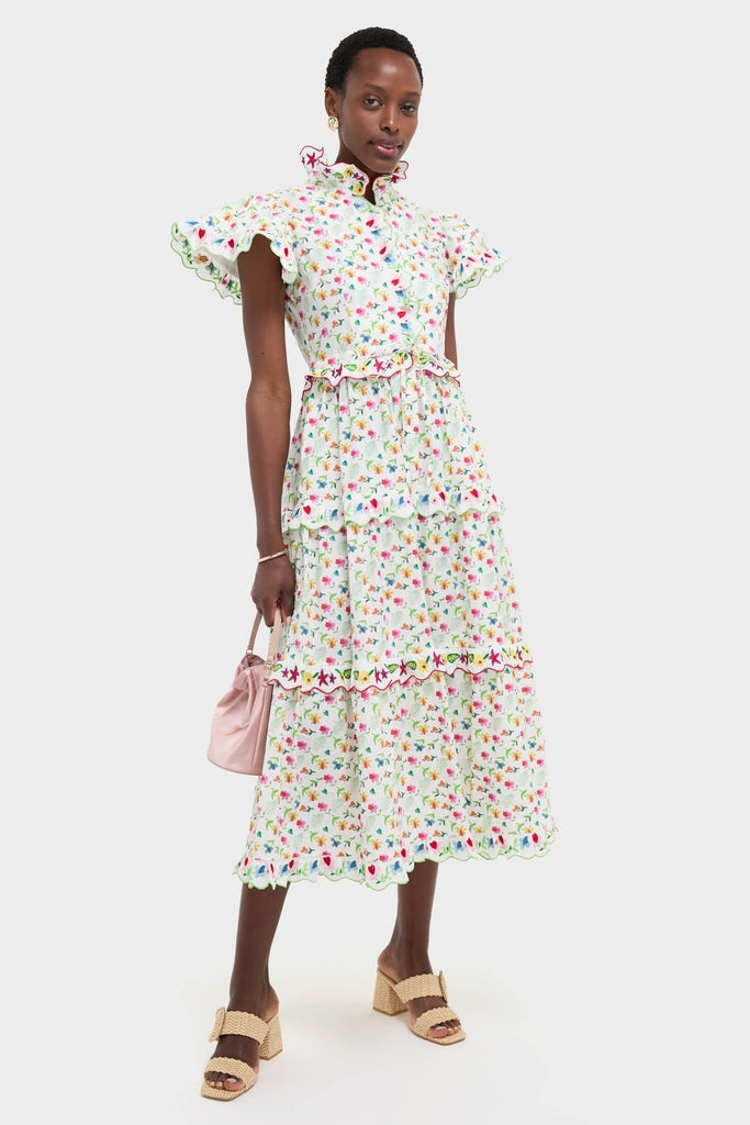 Woman models the White Nebula Dress, a classic, effortless calf-length floral dress with flutter sleeves, tiered scalloped skirt, paired with a pink pouch and block-heeled sandals.