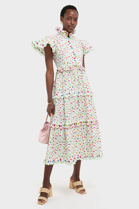 Woman models the White Nebula Dress, a classic, effortless calf-length floral dress with flutter sleeves, tiered scalloped skirt, paired with a pink pouch and block-heeled sandals.