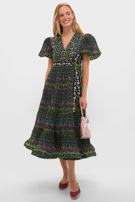 Woman in a classic, high-end Black Celestia Dress with multicolored floral prints, V-neckline, flared sleeves, and ruffled hem. Effortlessly styled with a pink bag and red pumps.