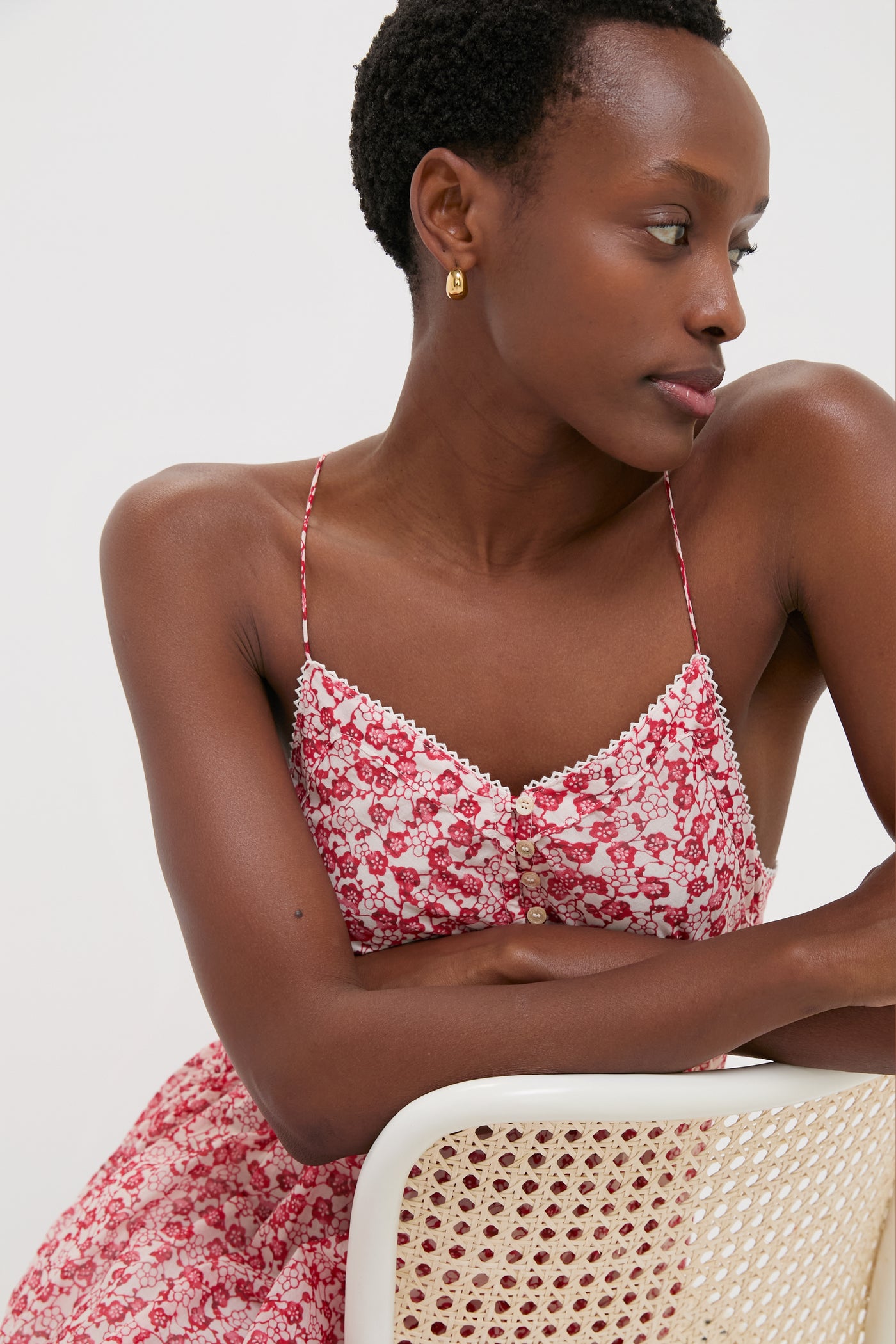 Young Black woman in Red Sabba Dress, classic and effortless with a tiered silhouette, spaghetti straps, and gold hoops, leaning on a woven chair, embodying high-end fun.