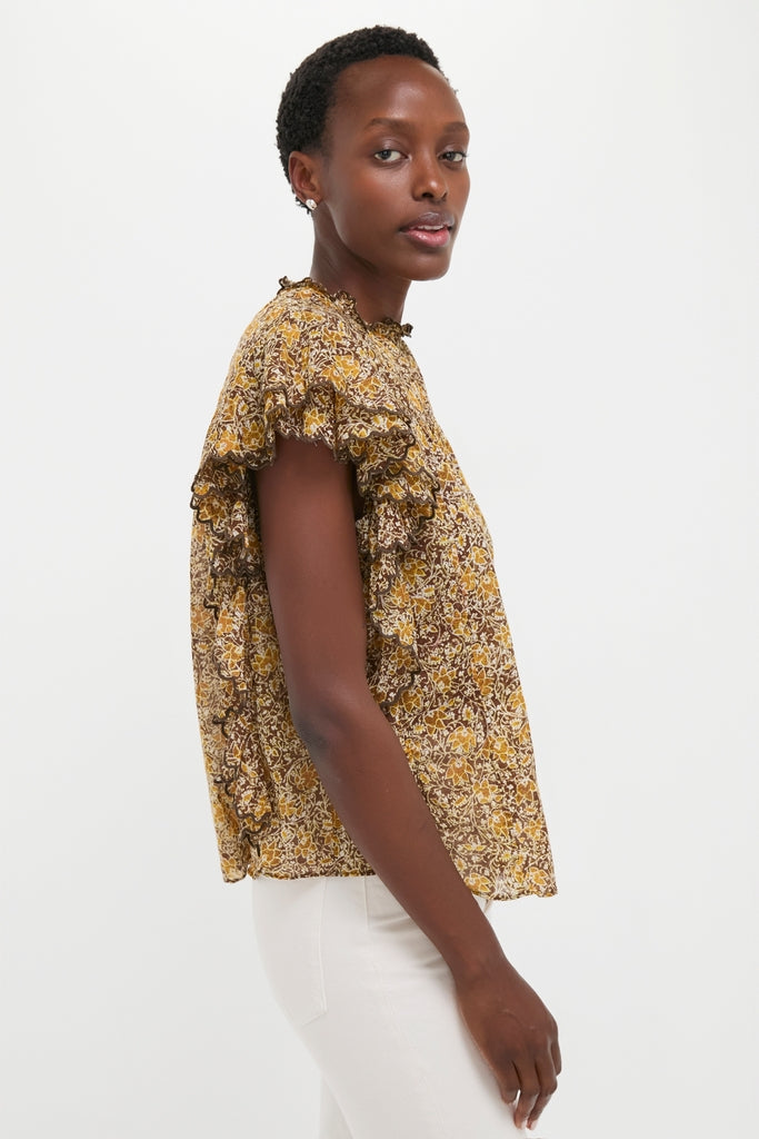 Model wearing the Khaki Daliyane Top, a classic, effortless blouse with a mustard-and-brown floral print and ruffle details, paired with white pants for a high-end, fun look.
