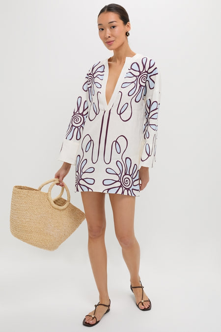 Woman models the Namur Celeste Siena Dress with floral motif, deep V-neck, and kimono sleeves, holding a straw tote; a classic, effortless resortwear choice.