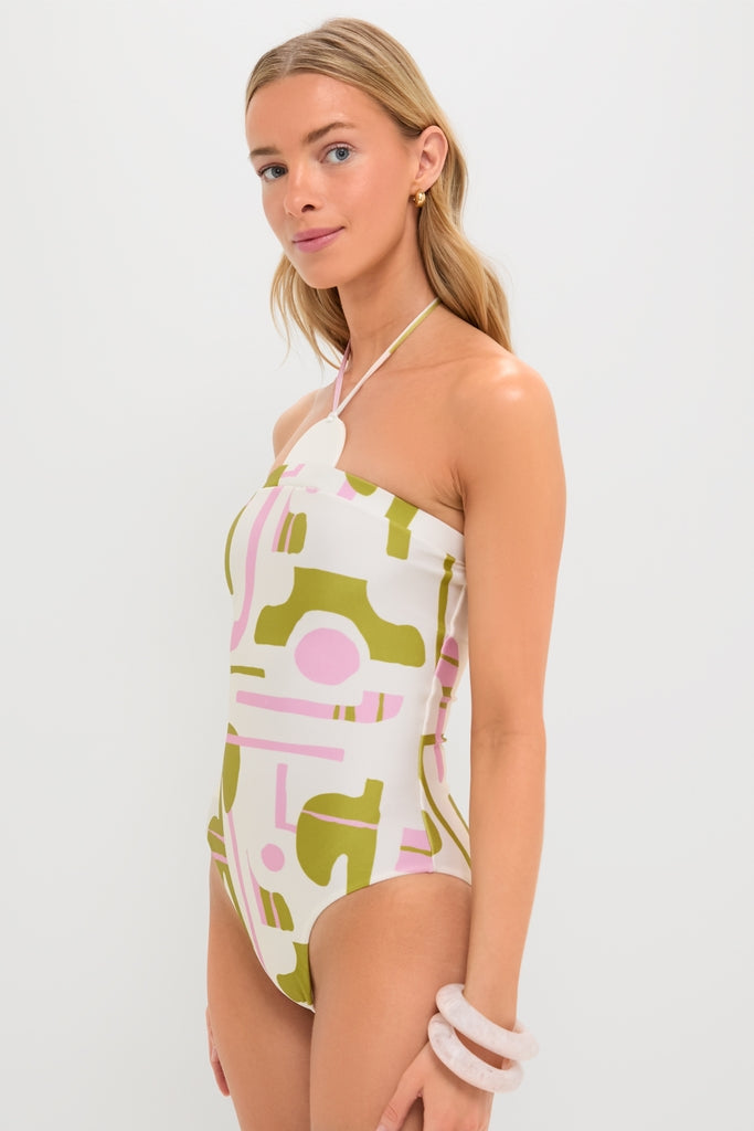 Woman models the Off-White Balearic Applique Swimsuit, featuring a classic halter neck and effortless olive-green and pale pink abstract shapes. She accessorizes with gold hoops and chunky bangles.