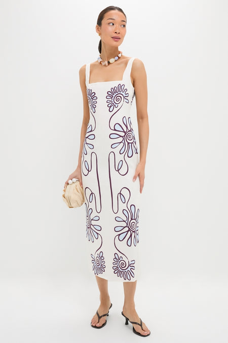 Woman in Namur Celeste Amaranta Dress, a classic, sleeveless white dress with purple swirls, accessorized with a chunky necklace and beige clutch, exuding effortless high-end style.