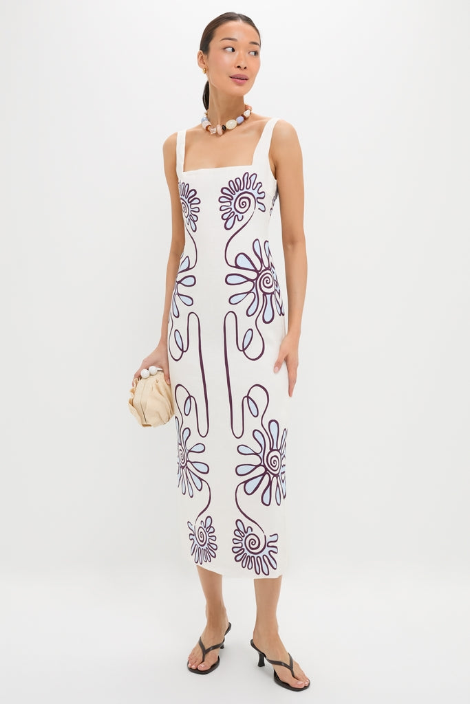 Woman in Namur Celeste Amaranta Dress, a classic, sleeveless white dress with purple swirls, accessorized with a chunky necklace and beige clutch, exuding effortless high-end style.
