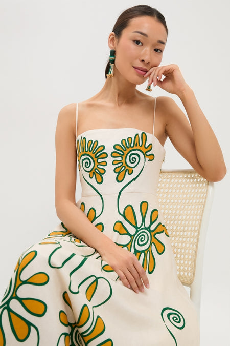 Woman in Namur Ivory Delta Dress with green and mustard floral motifs, seated on a cane-back chair, exuding an effortless, high-end, and fun style.