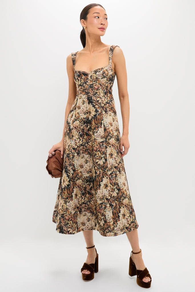 Woman models the Mirelle Jacquard Zinnia Midi Dress with earthy florals, featuring a square neckline and fitted bodice, paired with a brown clutch and platform heels.