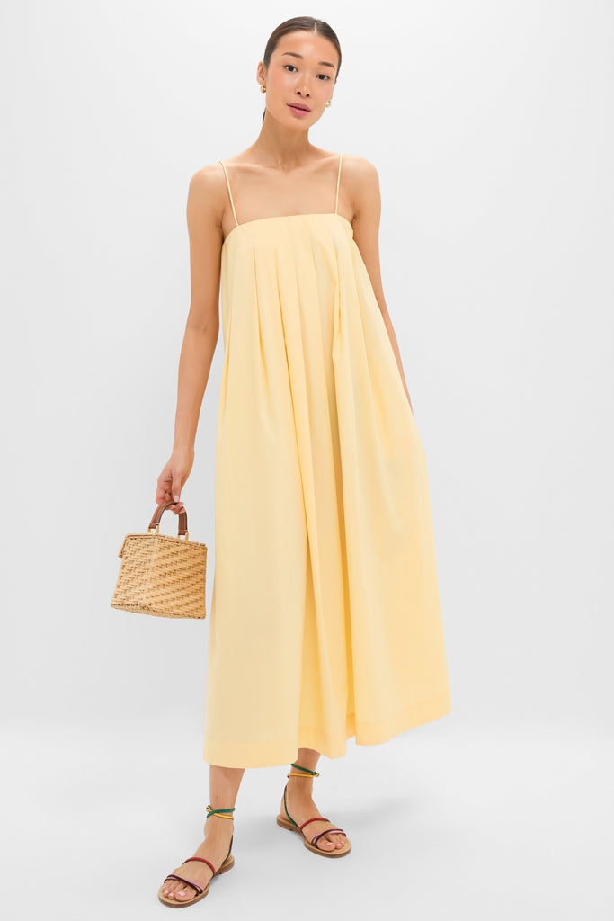 Model wearing classic Banana Lopez Midi Dress with adjustable straps and box pleats, styled effortlessly with a straw handbag and multicolored sandals for a high-end, fun look.