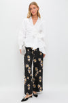 Model wearing classic Black and Yellow Daffy Trousers with scattered lilies, paired effortlessly with a white wrap blouse, mule heels, and a high-end silvery clutch.