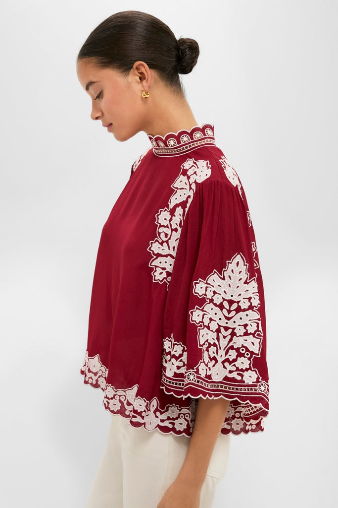 Woman in profile wearing the Burgundy Bicolor Palm Tree Embroidery Blouse, featuring a high-neck, loose fit, scalloped edges, and white floral details, paired with light pants for an effortless, classic look.