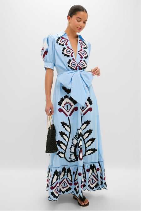 Woman modeling the Blue Artisanal Short Sleeve Maxi Dress—classic and effortless with a deep V-neck, puffed sleeves, and bold motifs—paired with a woven handbag and sandals.