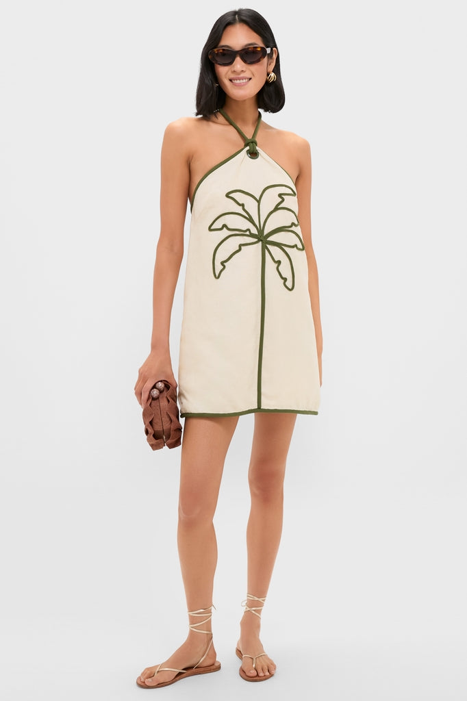Woman wearing the Sand Coconut Sleeveless Mini Dress with olive-green palm tree trim, accessorized with tortoiseshell sunglasses and a woven clutch, embodying classic, effortless, high-end style.
