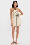Woman modeling the Sand Coconut Sleeveless Mini Dress, featuring a classic halter-neck, effortless natural canvas color, and fun palm tree detail, paired with strappy flat sandals.