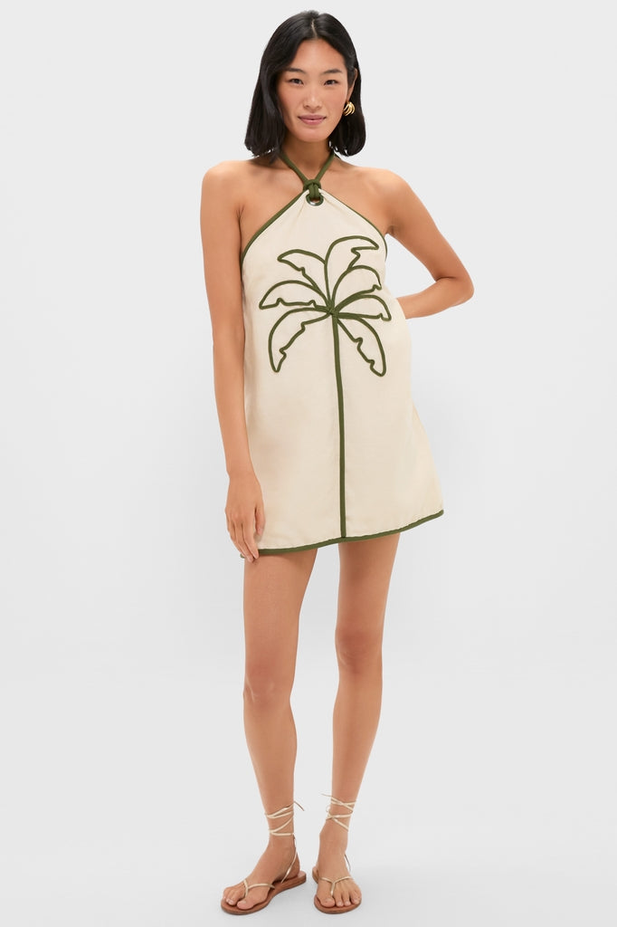 Woman modeling the Sand Coconut Sleeveless Mini Dress, featuring a classic halter-neck, effortless natural canvas color, and fun palm tree detail, paired with strappy flat sandals.