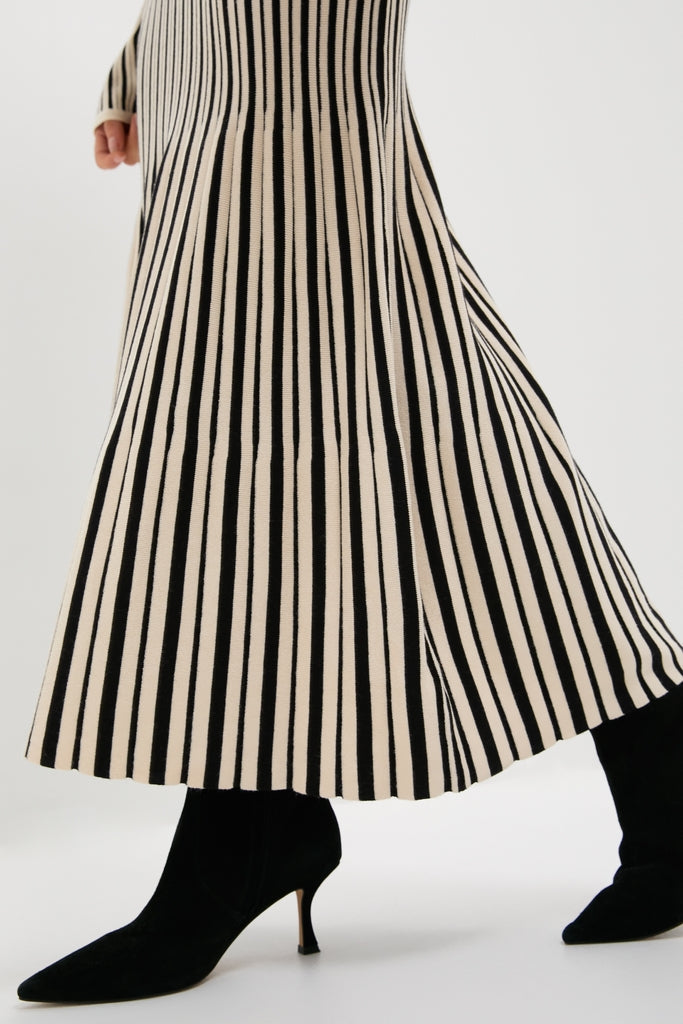 Person wearing the Black and Ivory Striped Maxi Dress with a flared hem and side slit, paired with classic black suede heeled boots for an effortless, high-end look.