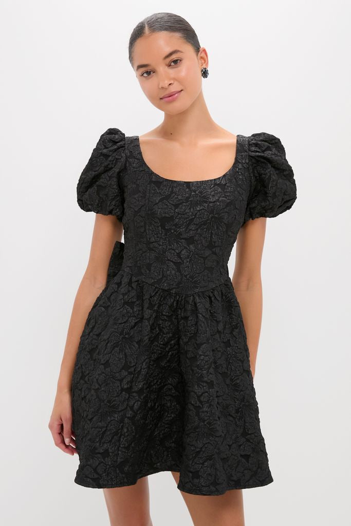 Young woman in a classic Black Jacquard Mini Dress featuring a textured floral pattern, scooped neckline, puffed sleeves, and flared skirt, embodying an effortless, high-end style.