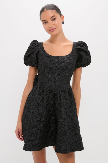 Young woman in a classic Black Jacquard Mini Dress featuring a textured floral pattern, scooped neckline, puffed sleeves, and flared skirt, embodying an effortless, high-end style.