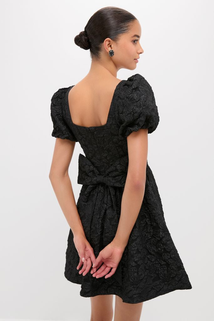 Young woman in a classic Black Jacquard Mini Dress with puffed sleeves, square back neckline, and waist bow, exuding effortless, high-end style.