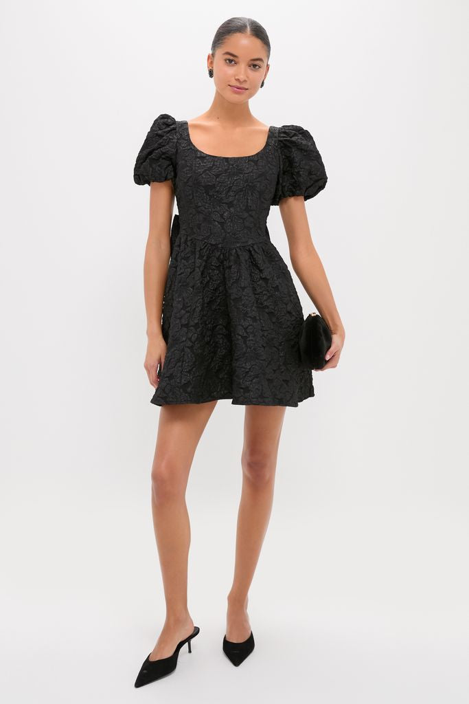 Model wearing a classic Black Jacquard Mini Dress with a square neckline and puffed sleeves, paired with a black clutch and pointed-toe mule heels, exuding effortless elegance.