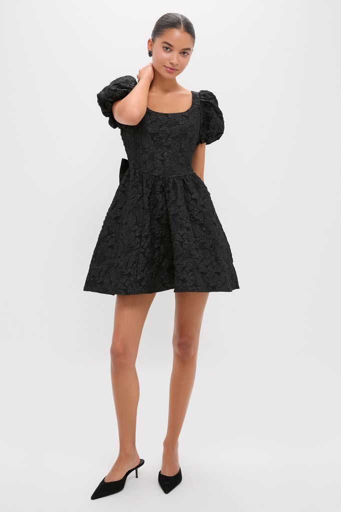 Young woman in a classic Black Jacquard Mini Dress with square neckline, puff sleeves, and flared skirt, embodying effortless elegance and fun, high-end style.