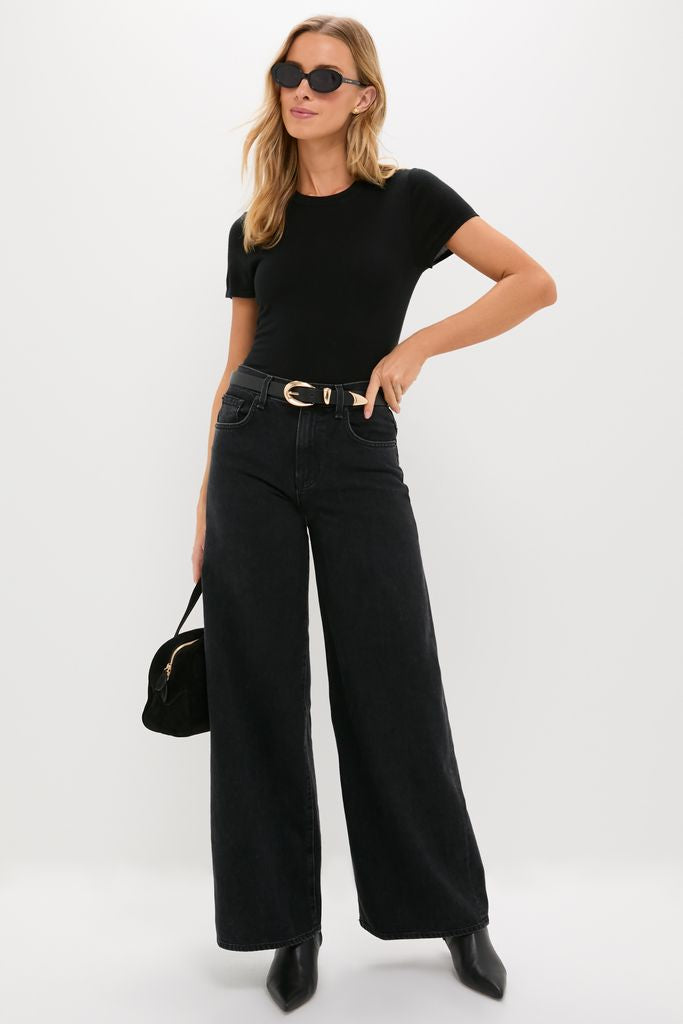 Woman wearing high-end Black Jeanne Sunglasses, striking a fashion pose. Her effortless style includes a fitted t-shirt, wide-leg jeans, and chic ankle boots.