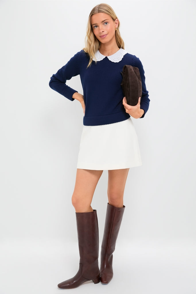 Young woman models the classic Navy Violette Sweater, featuring an eyelet lace collar, paired effortlessly with a white skirt and knee-high boots for a fun, high-end look.