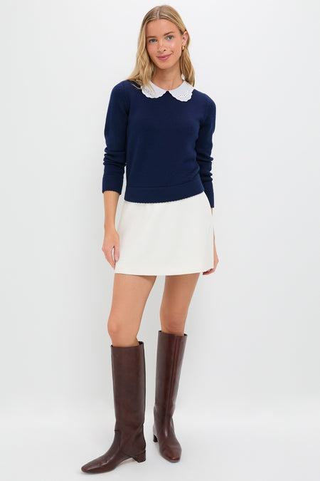 Woman models the classic Navy Violette Sweater with an eyelet Peter-Pan collar, paired effortlessly with a white skirt and high-end brown boots, embodying a fun, confident style.