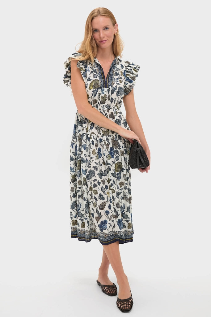Woman in classic white dress with blue and olive floral print, holding a clutch. Showcases Espresso Otto Hand-Knotted Flat Mule, embodying effortless, high-end style with fun woven design.