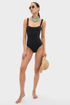 Woman models the classic Black Adelaide One Piece swimsuit with corset-style boning, chunky turquoise necklace, and aviator sunglasses, exuding effortless, high-end style.