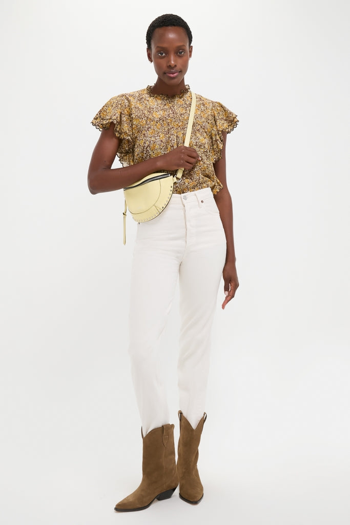 Woman wearing a classic, floral blouse with cream jeans and brown cowboy boots, showcasing the effortless Butter Skano Fannypack, a fun, high-end pale yellow crossbody bag.
