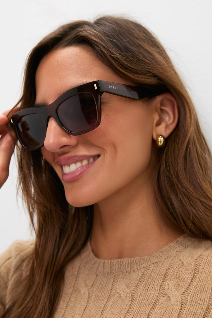 Woman wearing Whiskey Bradley Sunglasses by DIFF, featuring oversized dark square frames with gold accents, perfectly paired with a light brown sweater for a classic, effortless high-end look.