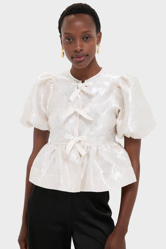 Egret Satin Cloque Peplum Blouse with puffed sleeves and decorative bows exudes a classic, effortless elegance, paired with dark high-waisted bottoms for a fun, high-end look.