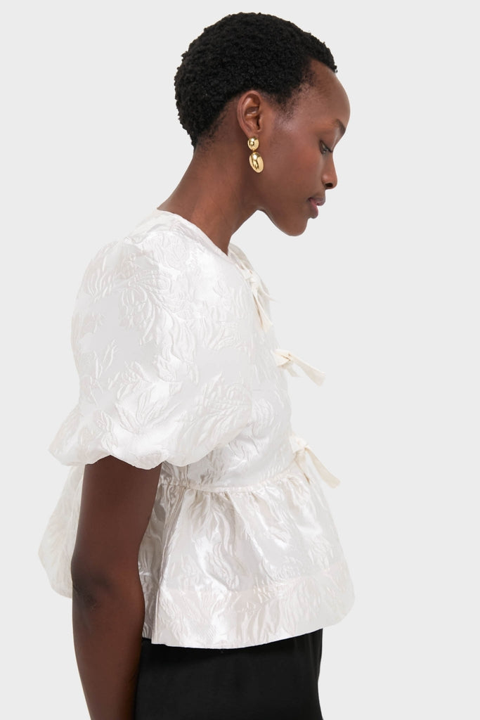 Egret Satin Cloque Peplum Blouse, featuring a textured, jacquard-style design with bow-tie closures, worn by a woman exuding classic, effortless elegance and fun, high-end style.
