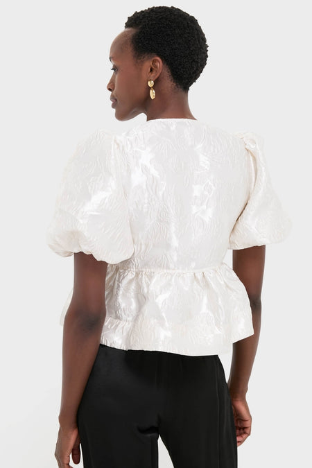 Person wearing the classic Egret Satin Cloque Peplum Blouse with puffed sleeves, embodying an effortless, high-end style, paired with black trousers and gold drop earrings.