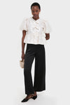 Woman in Egret Satin Cloque Peplum Blouse with puffed sleeves and decorative bows, paired with classic high-waisted trousers and slingback flats, exuding effortless, high-end style.