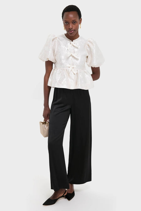Woman in Egret Satin Cloque Peplum Blouse with puffed sleeves and decorative bows, paired with classic high-waisted trousers and slingback flats, exuding effortless, high-end style.