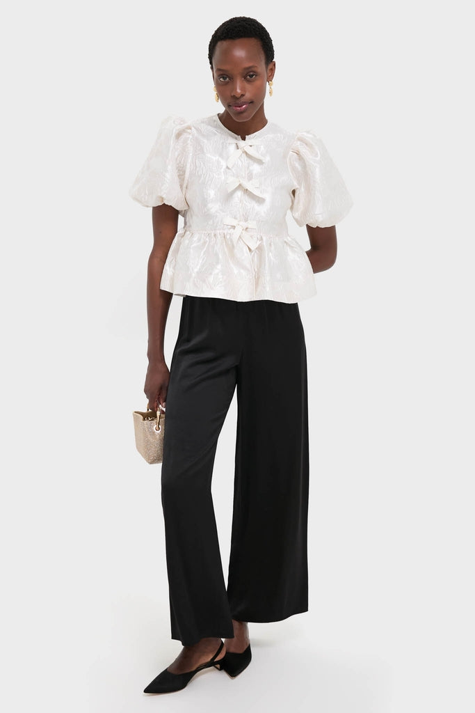 Woman in Egret Satin Cloque Peplum Blouse with puffed sleeves and decorative bows, paired with classic high-waisted trousers and slingback flats, exuding effortless, high-end style.
