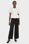 Woman wearing the Egret Satin Cloque Peplum Blouse with jacquard texture, puff sleeves, decorative bows, paired with black wide-leg trousers and slingback heels, epitomizing effortless, high-end style.