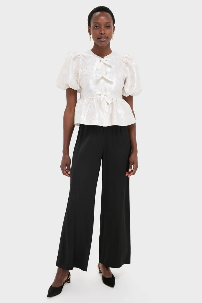 Woman wearing the Egret Satin Cloque Peplum Blouse with jacquard texture, puff sleeves, decorative bows, paired with black wide-leg trousers and slingback heels, epitomizing effortless, high-end style.