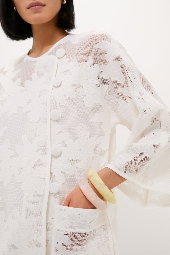 White Floral Lace Sidney Coverup: Classic, sheer knit cardigan with floral lace, fabric-covered buttons, patch pocket, and wide sleeves, exuding effortless, high-end style. Perfect for fun, confident looks.